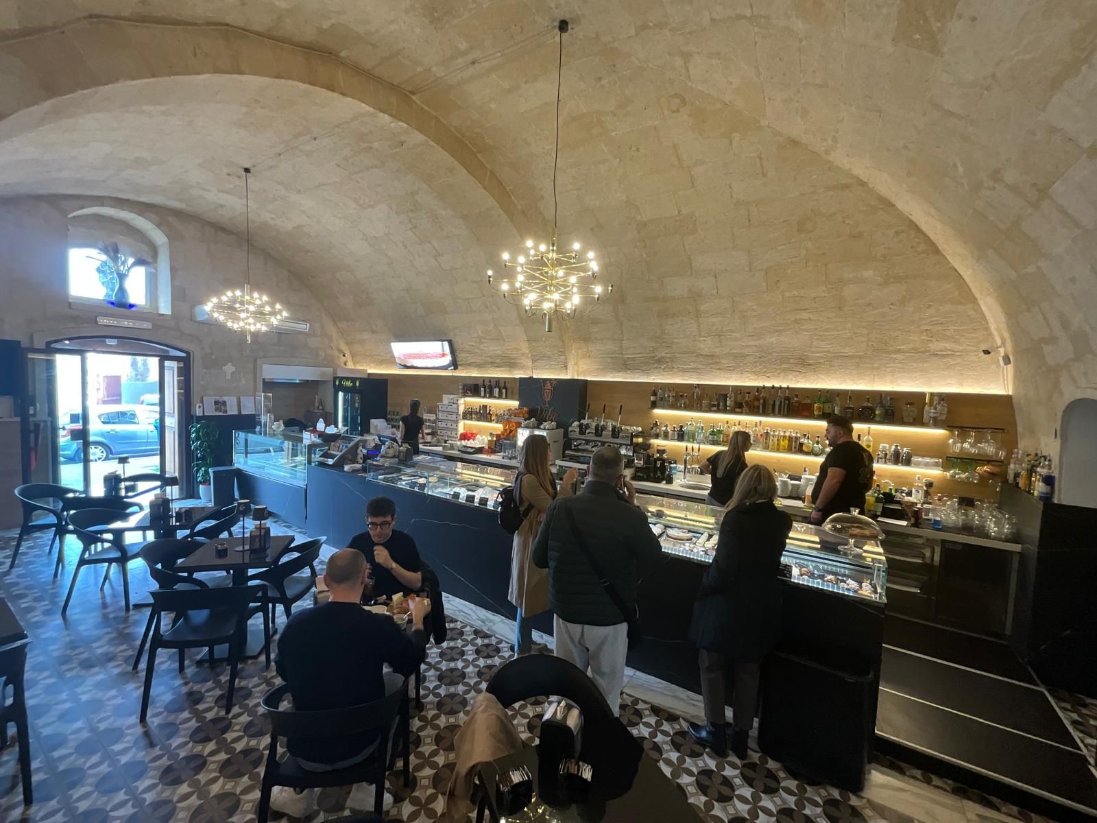 L'interno di una moderna panetteria presenta un soffitto a volta in pietra, clienti seduti ai tavoli e un bancone di servizio con vetrine espositive.