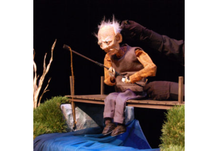 Puppet figure sitting on a small wooden bridge over blue water, holding a stringed instrument at night