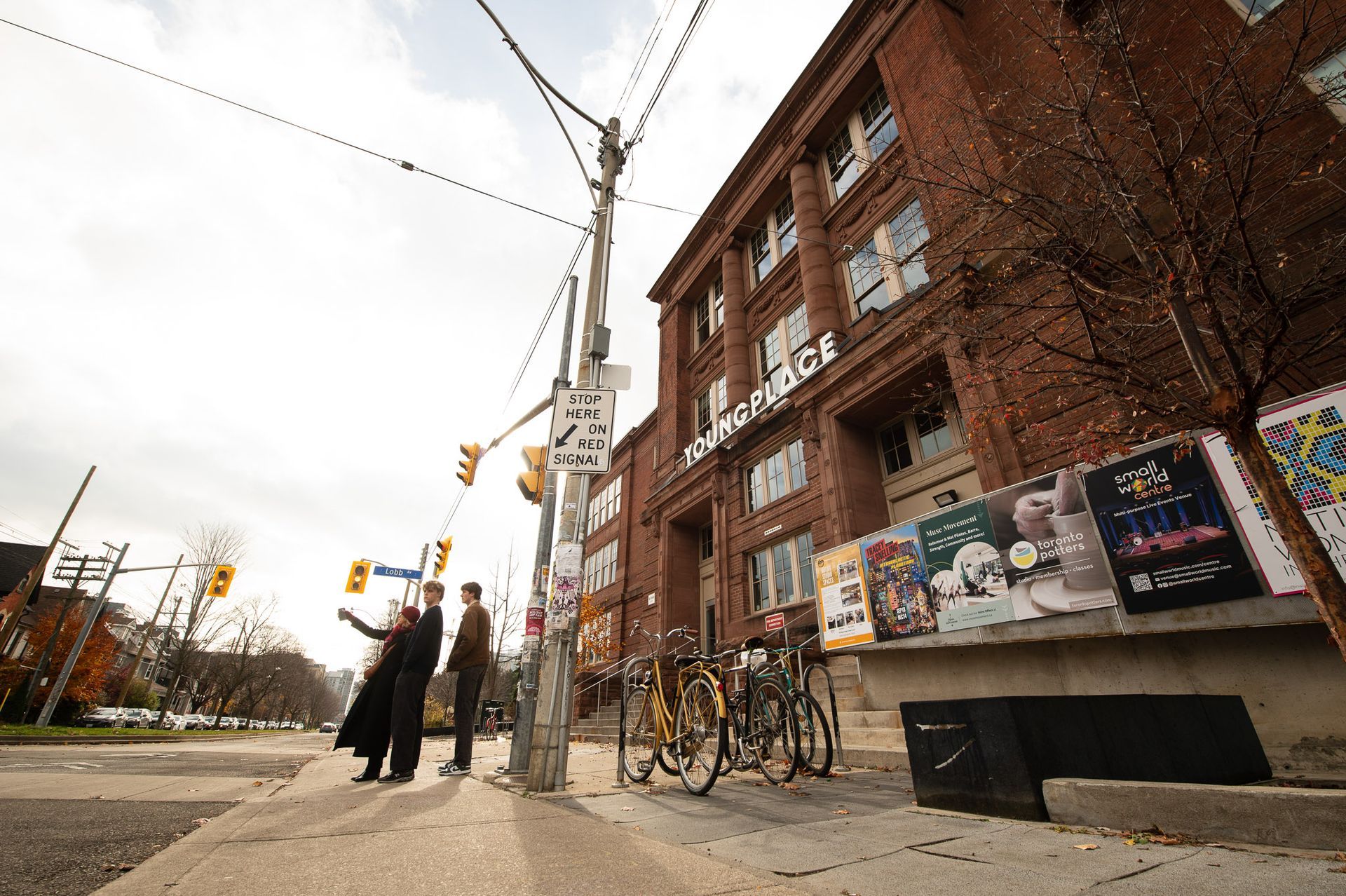 Two people on a city sidewalk beside a brick building, bicycles, and a street sign at an intersection.