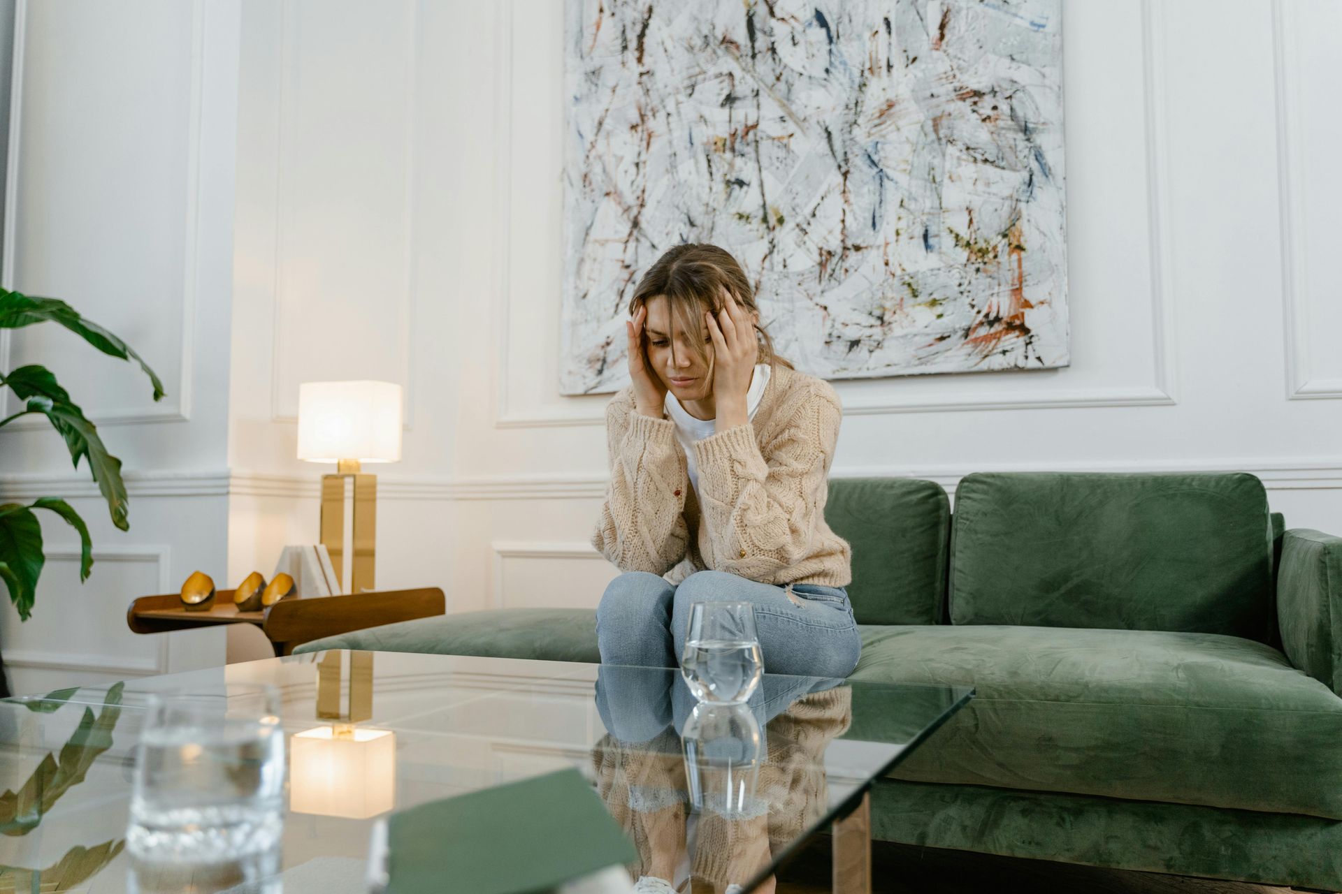 Woman with hands on head, seated on a green sofa, looking distressed. White room, abstract art, glass of water.