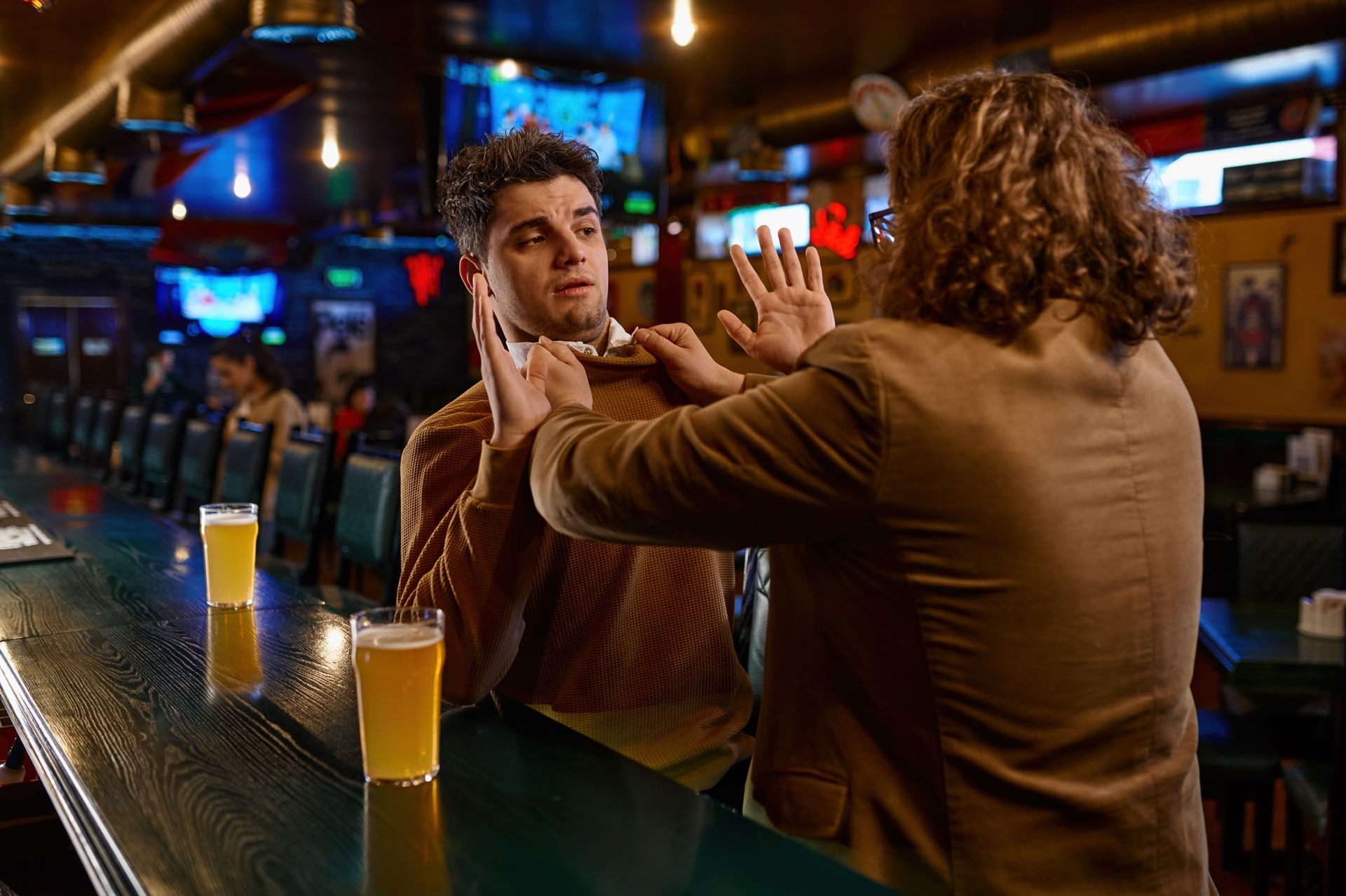 Two men arguing in a bar. One man is being grabbed by the other's hands. Two beers sit on the bar.
