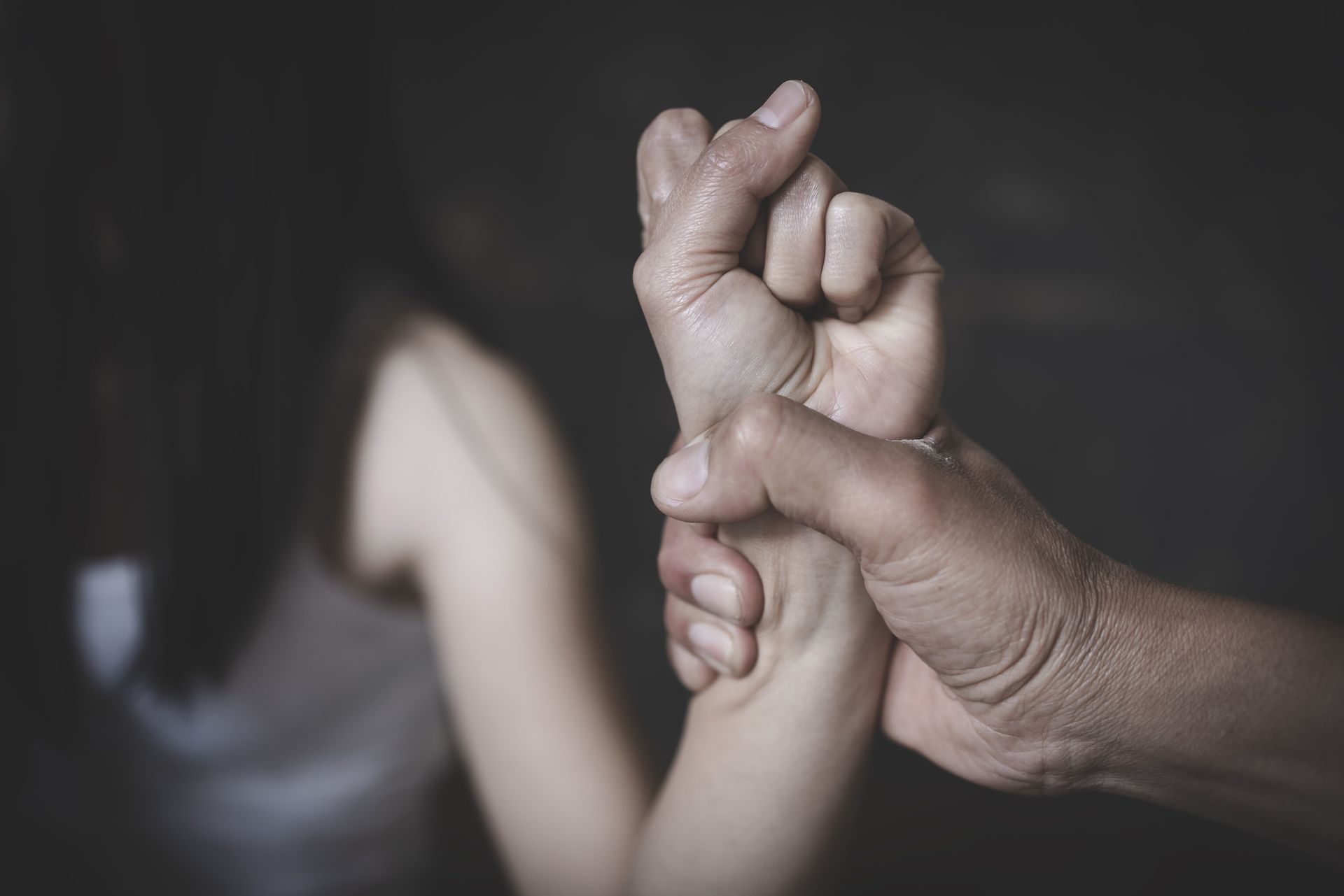 Person's hand gripping another's wrist. In background, blurred figure of a person in a distressed posture.