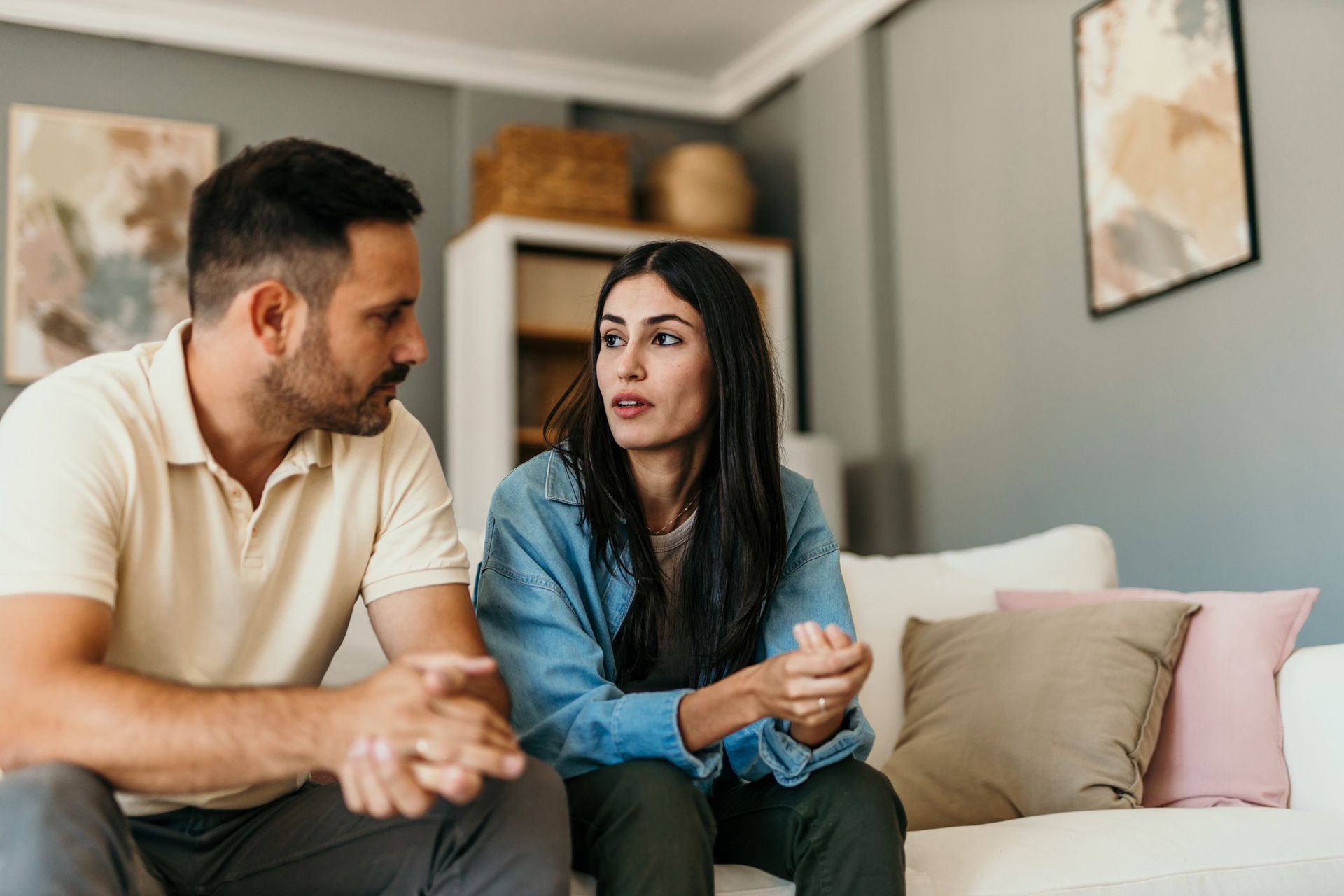 A man and woman sitting on a couch, talking. The woman is gesturing with her hands, looking concerned.