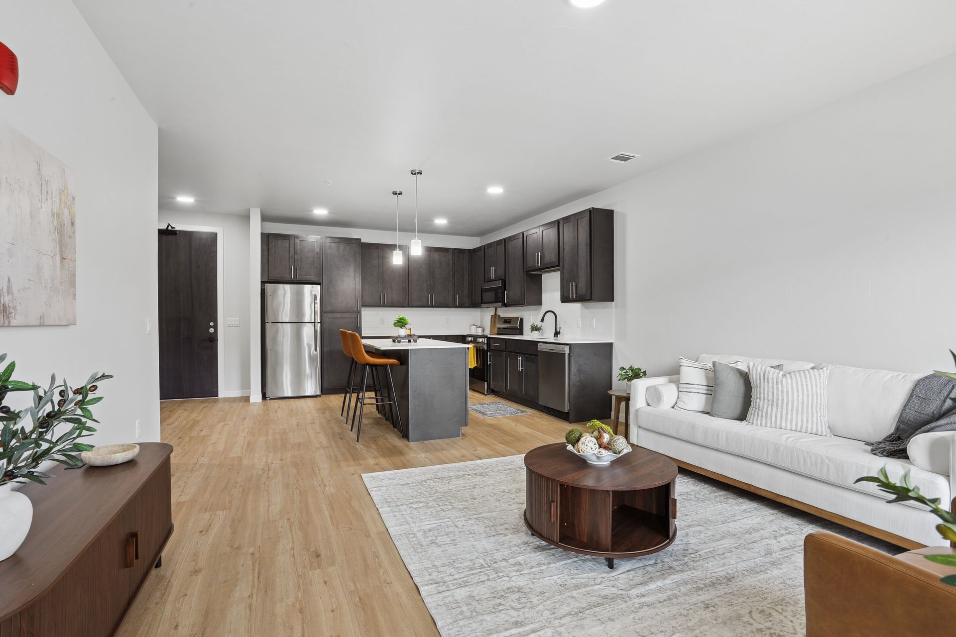 Open-plan living room and kitchen with wood floors, dark cabinets, white sofa, and gray island stools