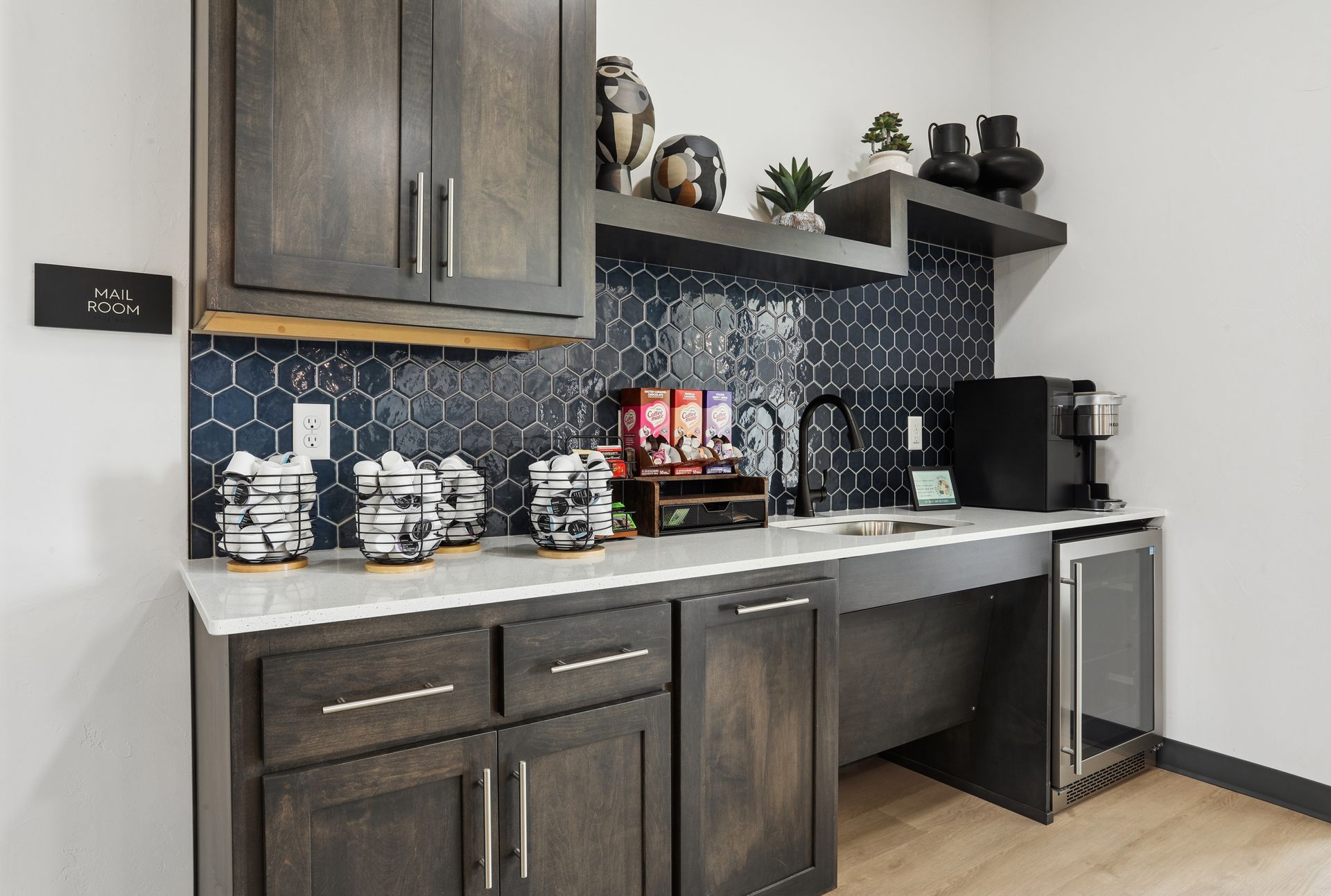 Coffee station with dark cabinets, white countertop, navy tile backsplash, and coffee maker on the right