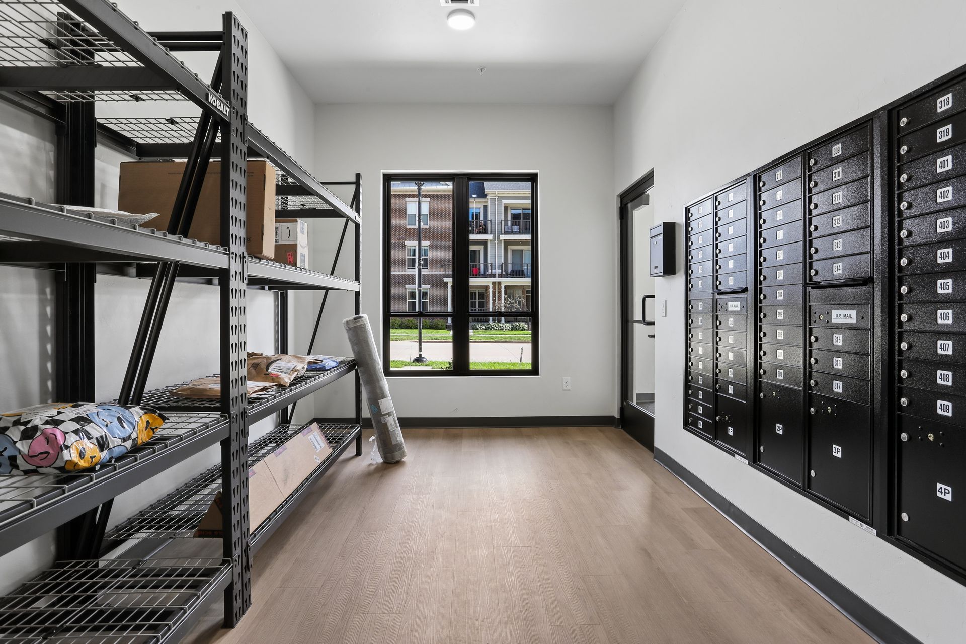 Modern apartment mailroom with black parcel lockers and metal shelving, viewed through a window.