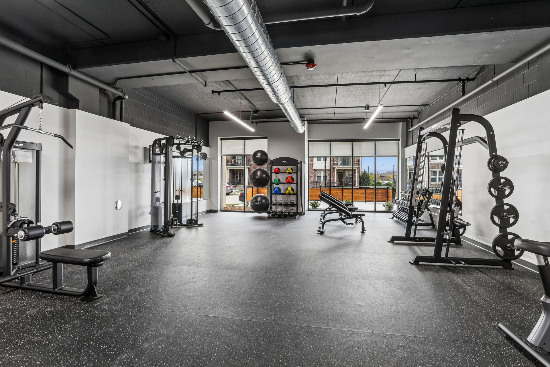 Modern gym with weight machines, benches, and black rubber flooring under exposed ceiling beams.