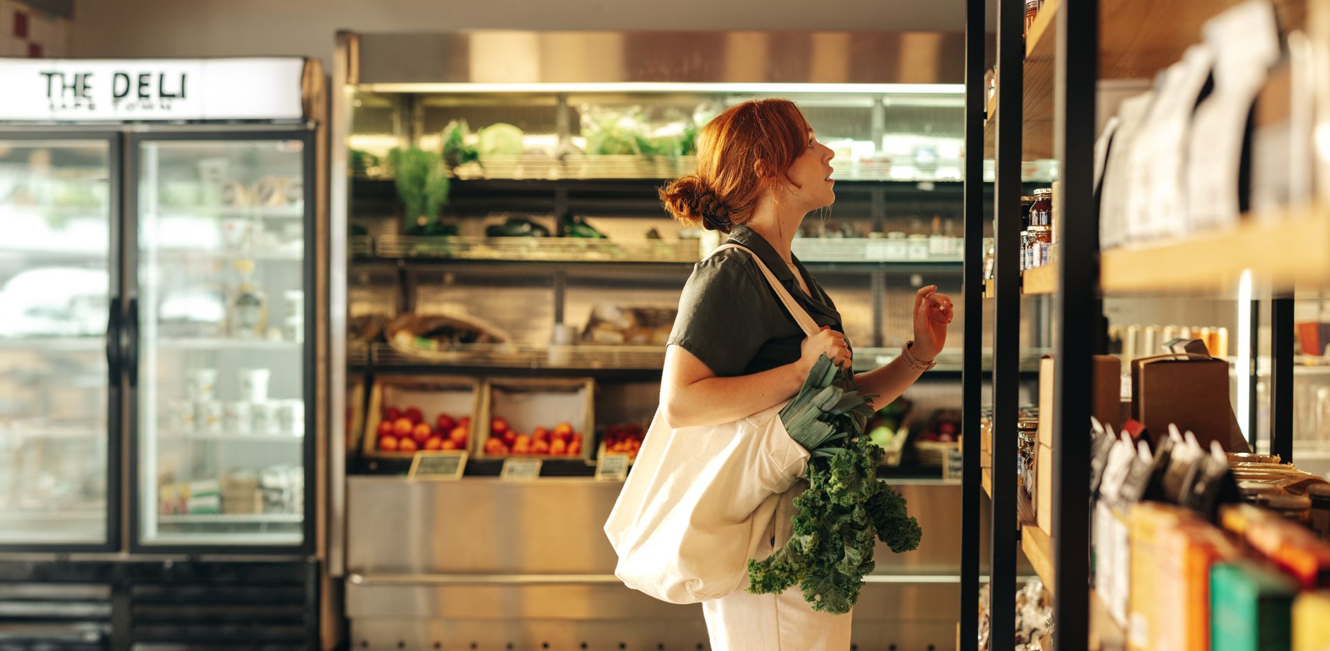 A side view of a young woman with red hair pulled back in a bun, browsing the shelves of a modern grocery store or deli market.