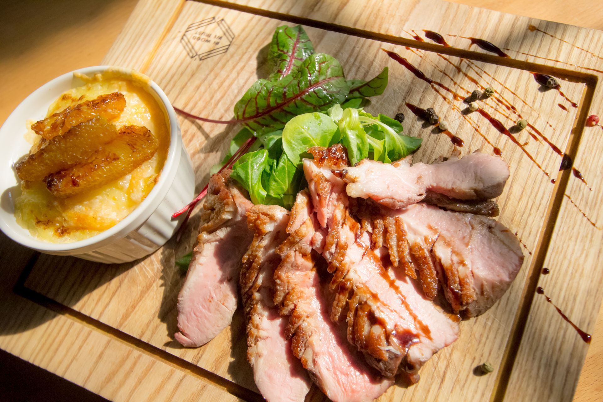 A top-down, close-up shot of sliced, tender roast meat with a balsamic drizzle served on a light wooden board. The dish is garnished with fresh green salad leaves and red-veined sorrel, alongside a small white ramekin containing a side dish under bright, direct sunlight.