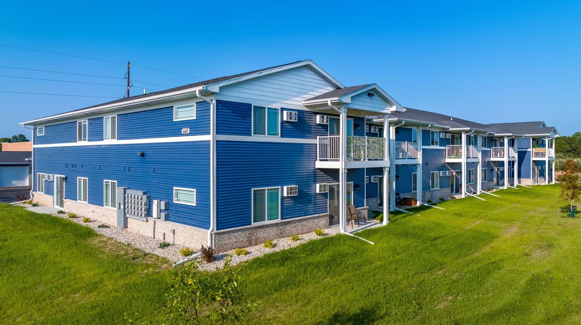 Blue coastal apartment building with balconies and green lawn under a clear sky