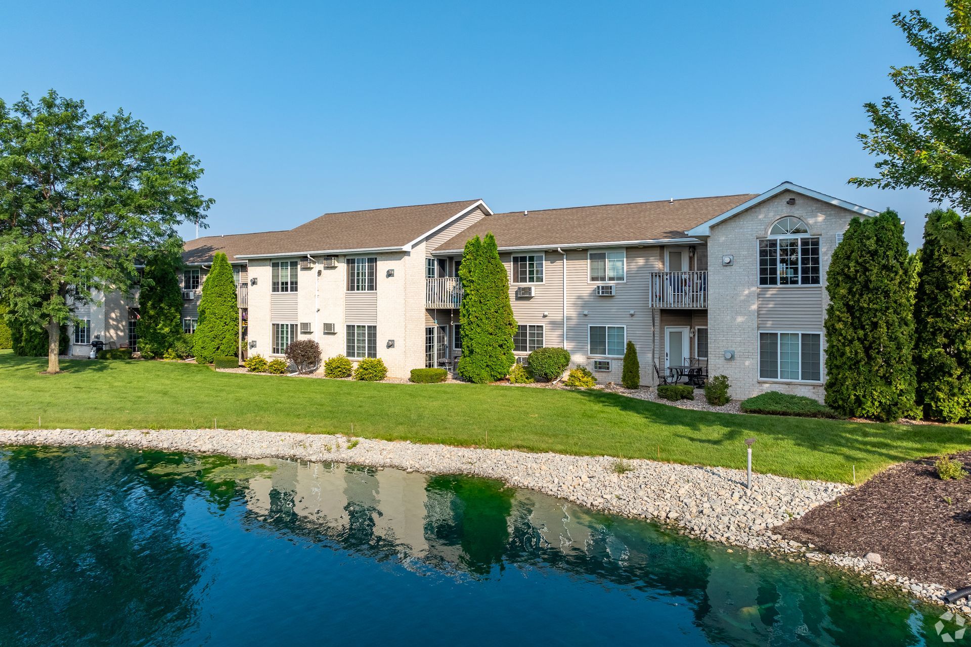 Apartment building beside a pond with green lawn and trees on a sunny day