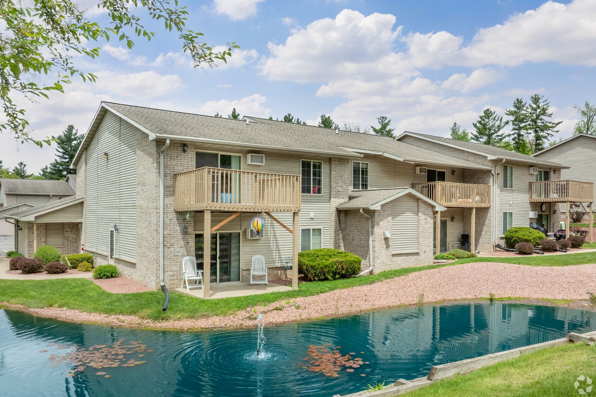 Apartment complex with balconies beside a blue pond and green landscaping under a cloudy sky