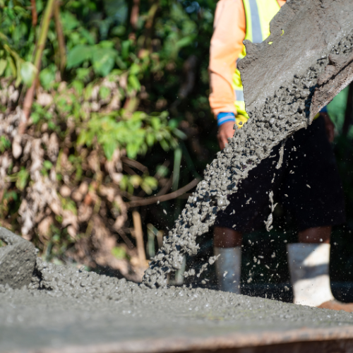 Worker pouring wet concrete, wearing safety vest, onto a surface; greenery in background.