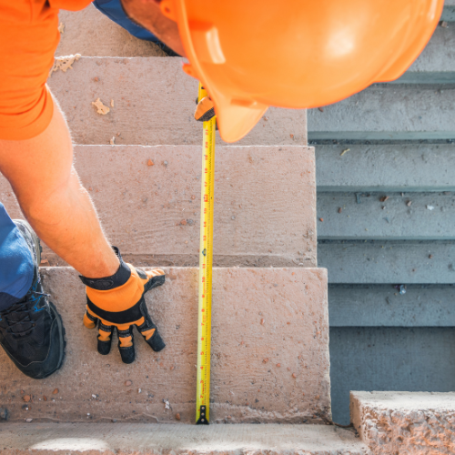 Construction worker measures a concrete stair with a yellow tape measure, wearing a hard hat and work gloves.