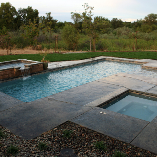Pool with a waterfall feature and adjacent spa, set against a grassy lawn and trees.