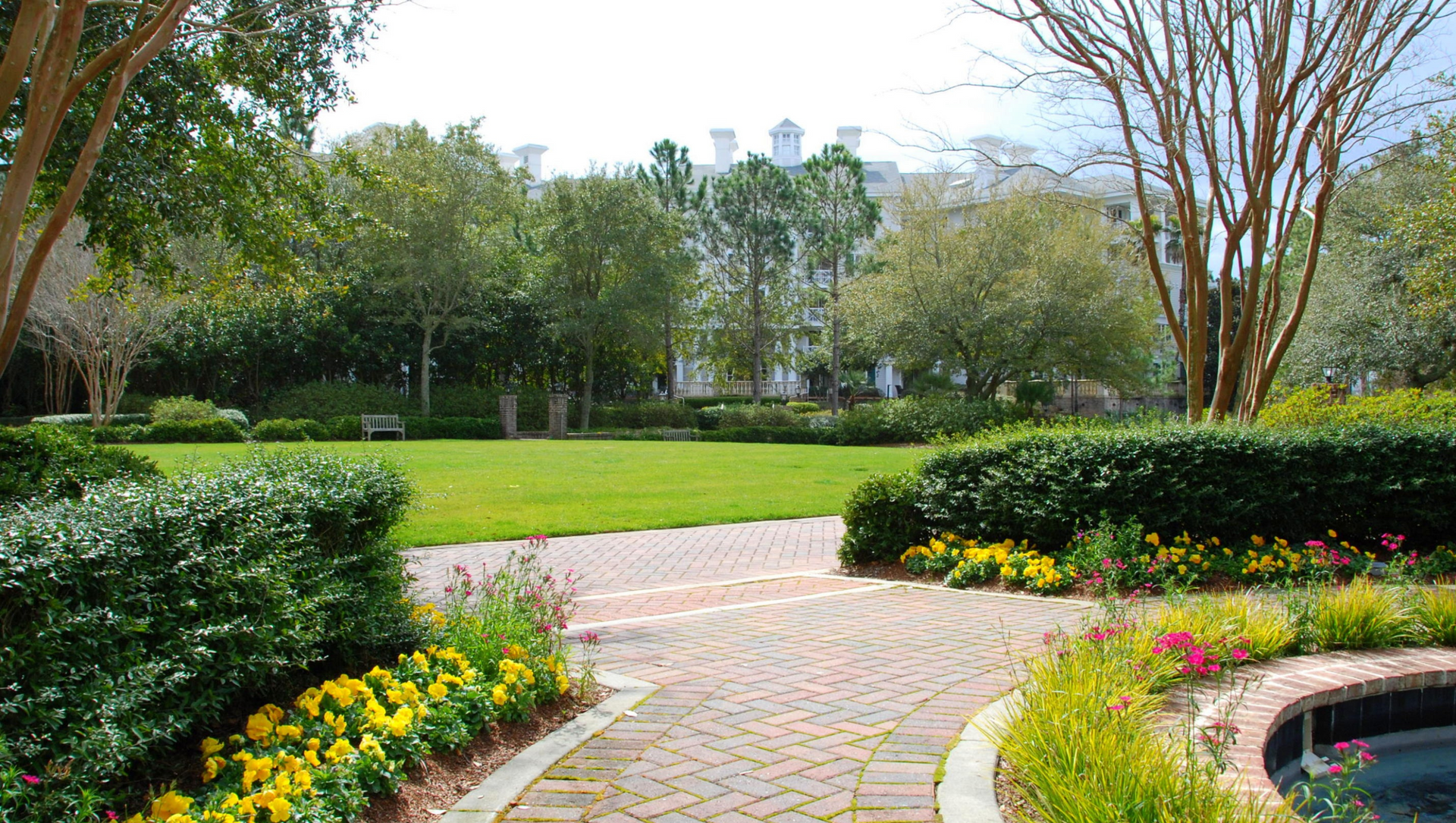 Brick pathway through a lush garden with a grassy lawn and trees, a large white building in the background.