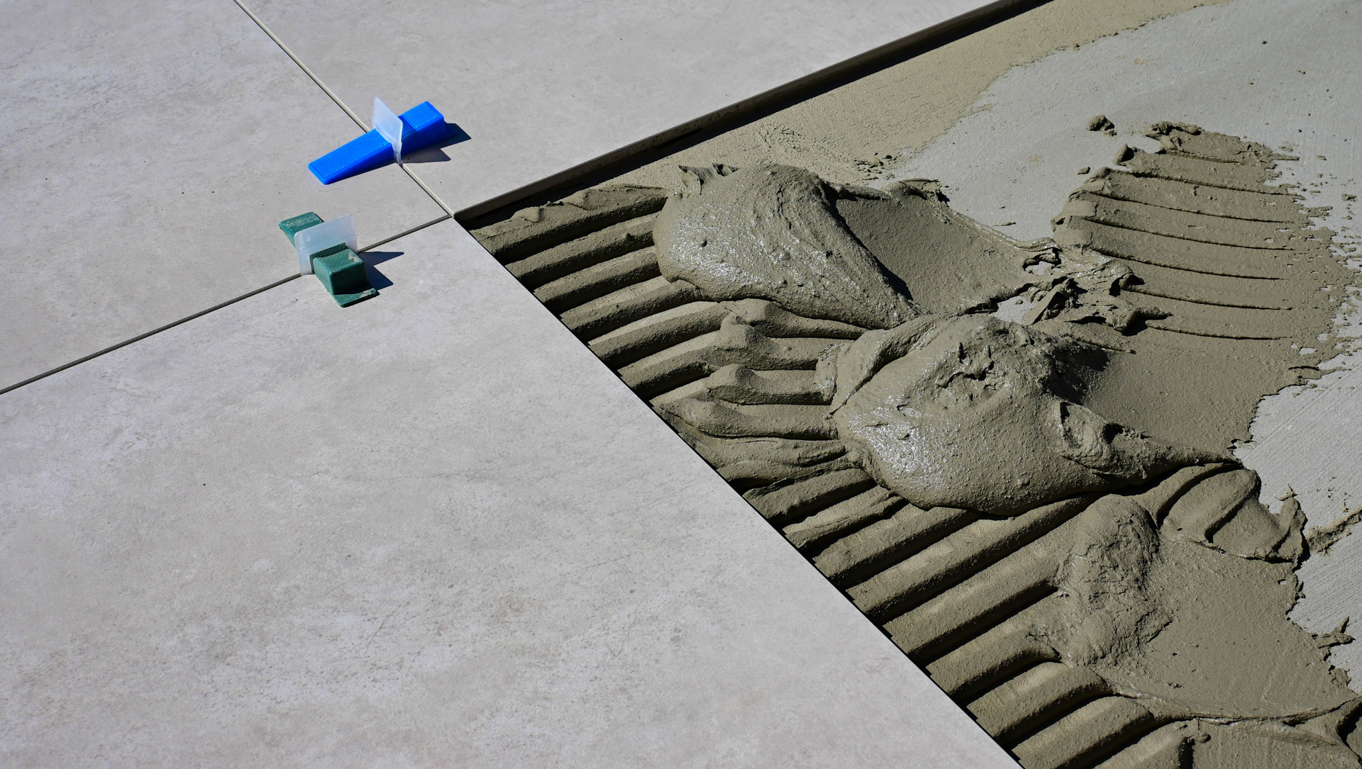 Laying floor tiles with notched trowel, mortar, and leveling clips. Light gray tiles on concrete.