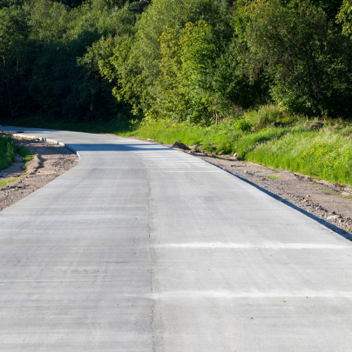 Newly paved gray concrete road winding through a green wooded area.