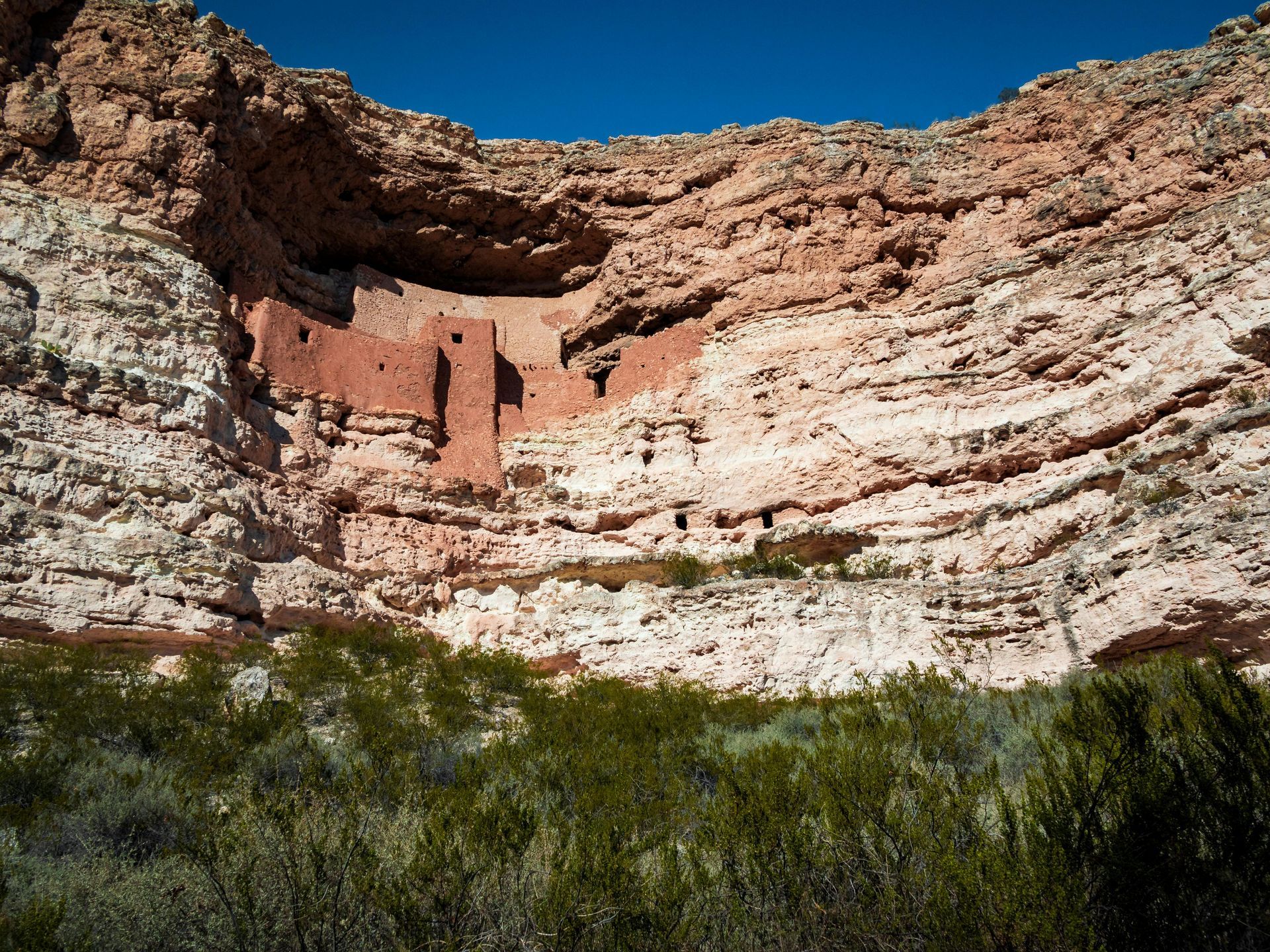 Cliff dwellings carved into a layered red rock canyon with green vegetation in the foreground.