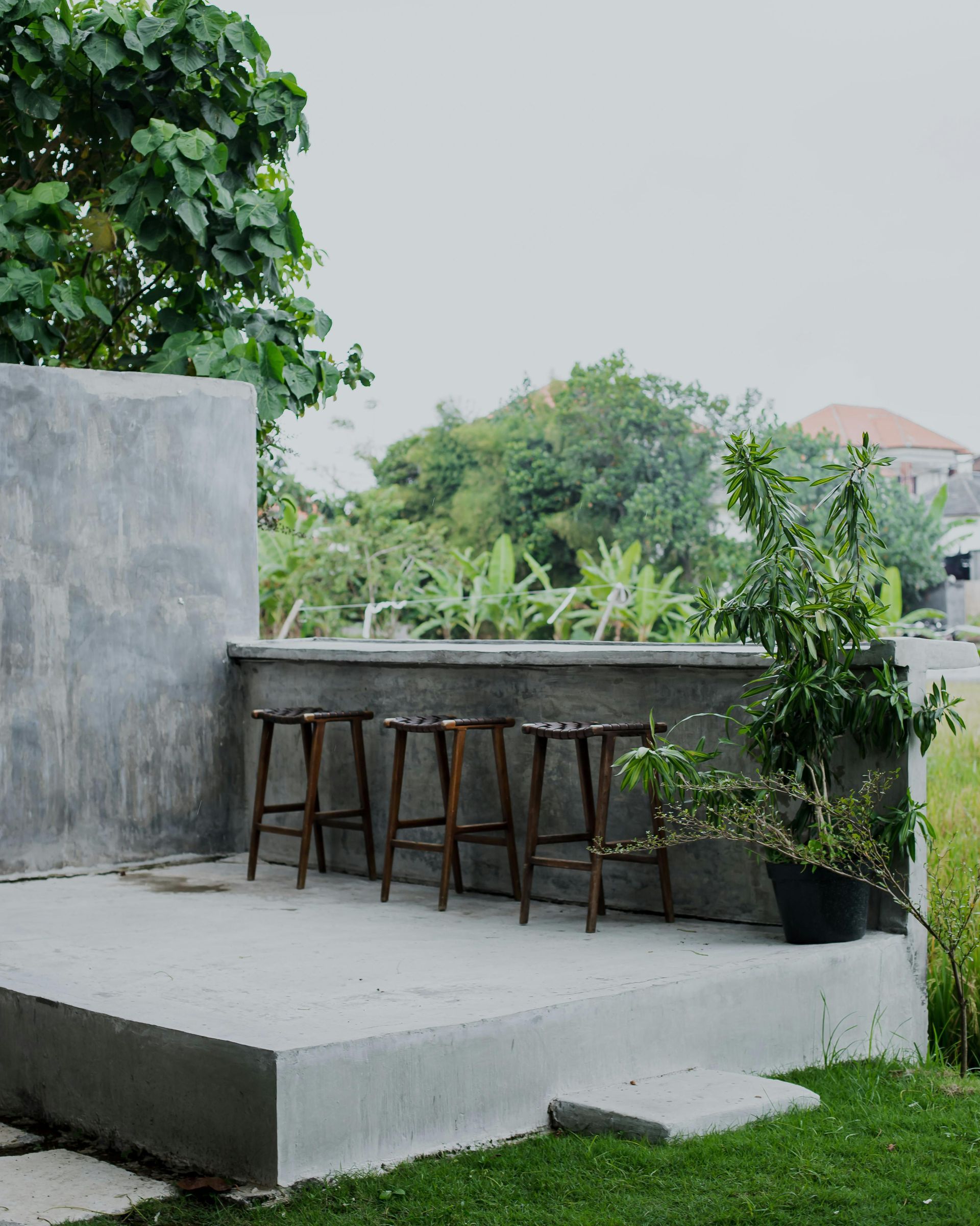 Outdoor concrete bar with four wooden stools, greenery backdrop.