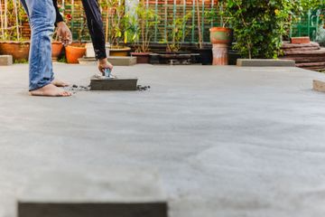 Person smoothing wet concrete with a trowel on an outdoor patio, surrounded by plants in pots.