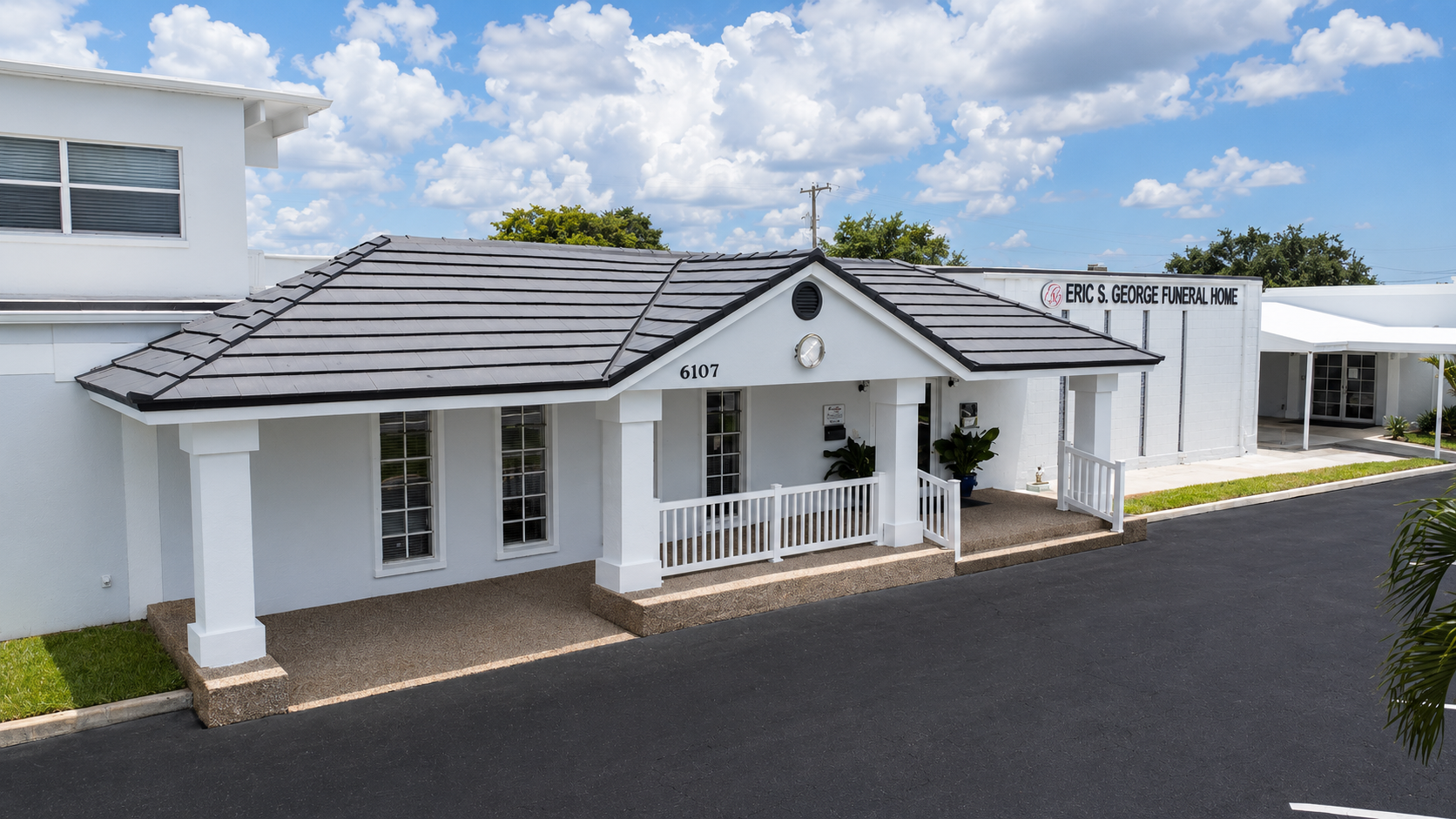 White house with porch and dark roof beside a paved driveway under a partly cloudy sky