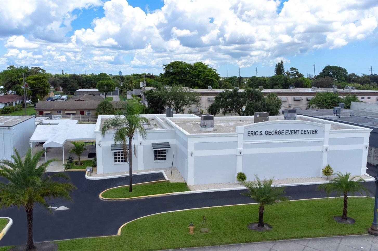 White school building with palm trees and lawn, viewed from above under a partly cloudy sky
