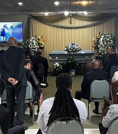 Funeral service with a casket, floral sprays, and seated mourners in a chapel.