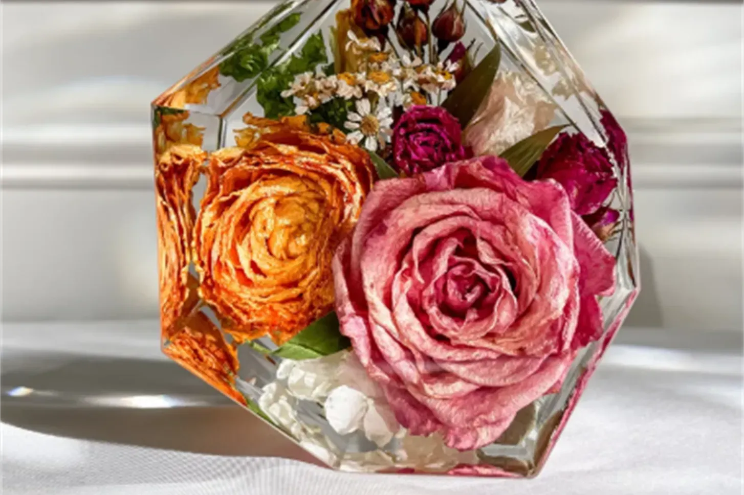 Glass vase with orange and pink roses, white flowers, and greenery on a sunlit surface