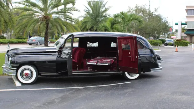 Black vintage hearse with open side doors, showing red interior, parked in a lot with palm trees.