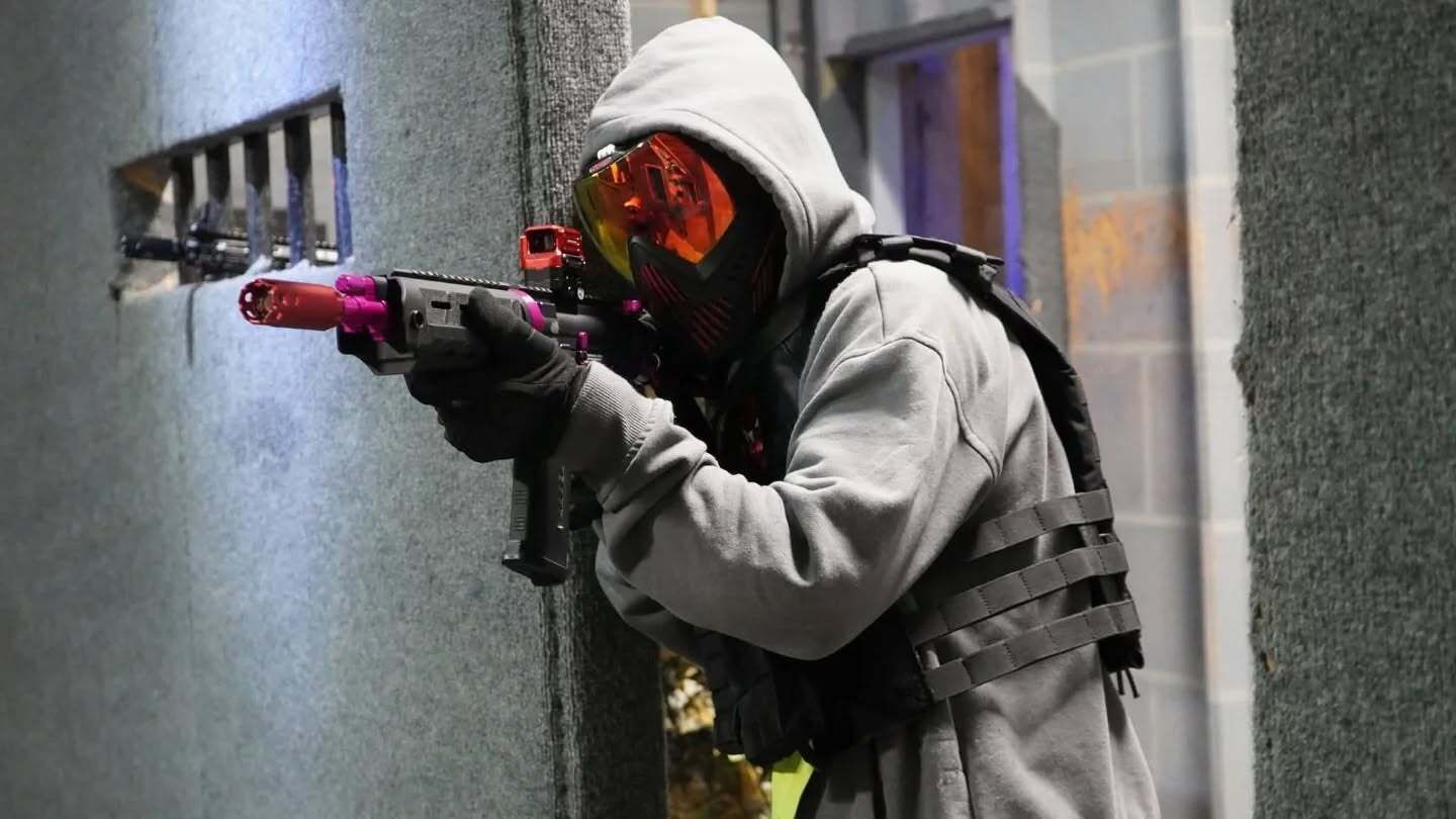 Person in tactical gear aiming a paintball gun. They wear a hood and mask, standing near a building.