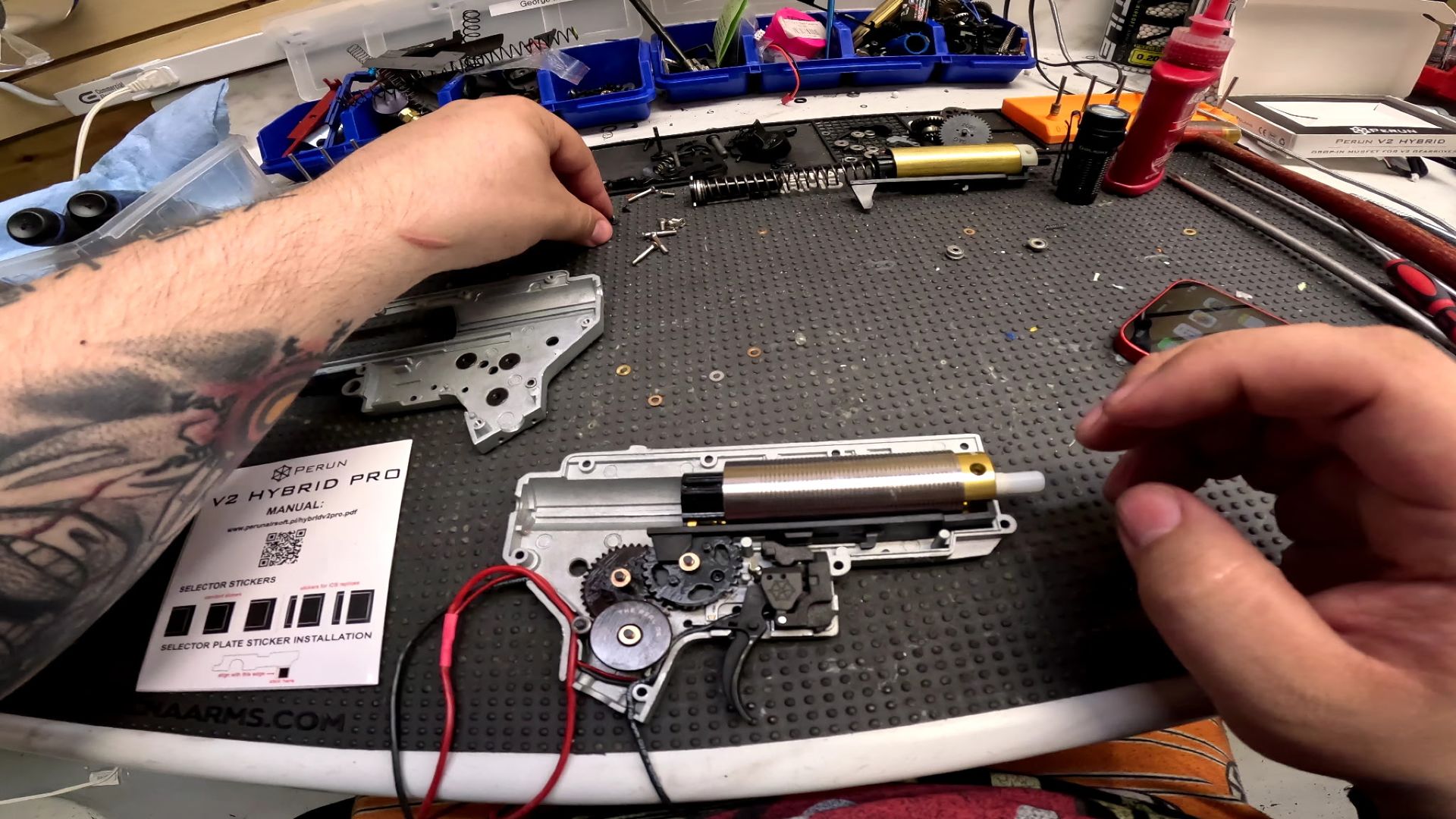 Hands assembling an airsoft gun on a workbench with parts scattered around.
