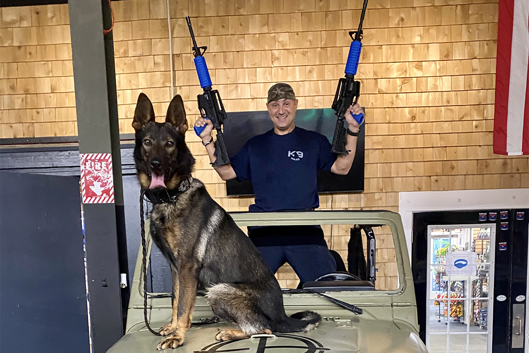 A man holding blue and black rifles, behind a jeep with a German Shepherd. American flag in the background.