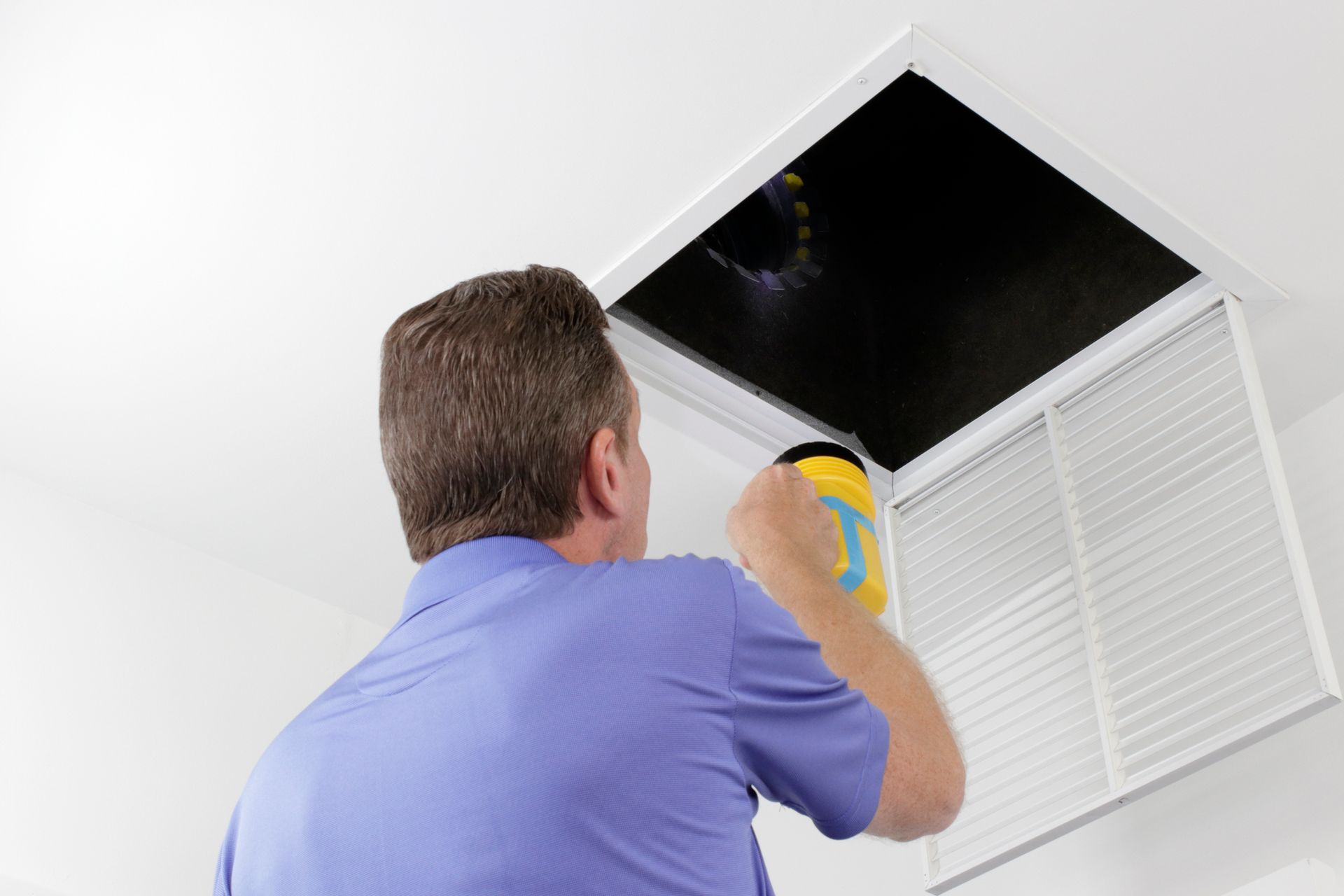 HVAC technician inspecting ceiling air vent with flashlight.