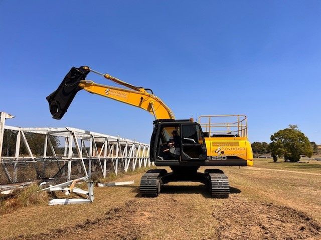 Yellow excavator demolishing a white fence on a sunny day, with a blue sky and field in the background.