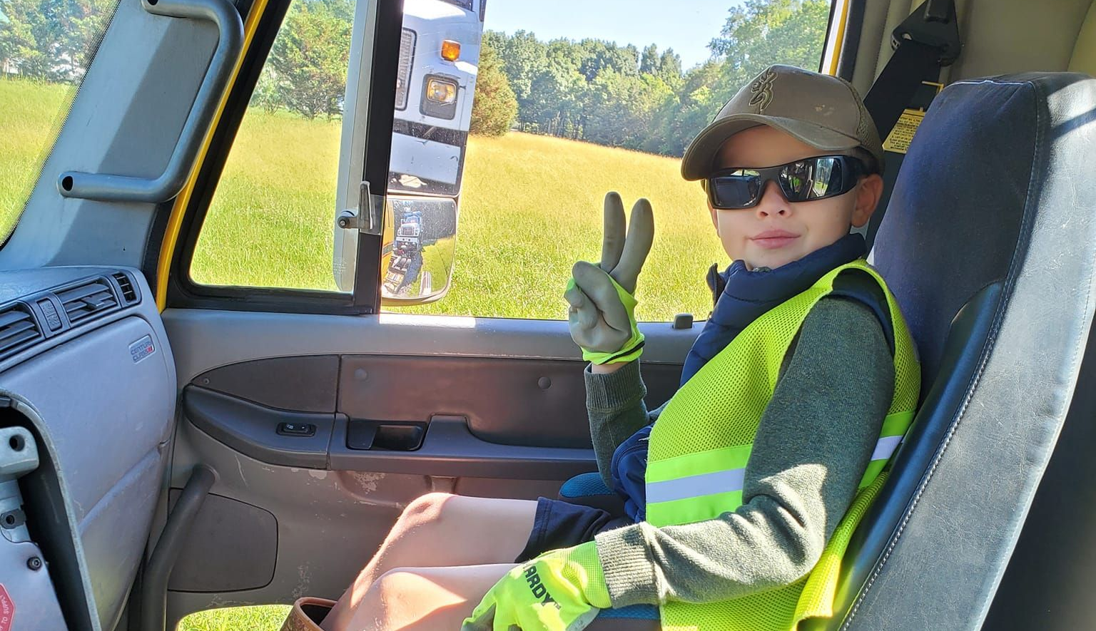 A person wearing a high-visibility vest, cap, and sunglasses sits inside a vehicle, making a peace sign toward the camera.