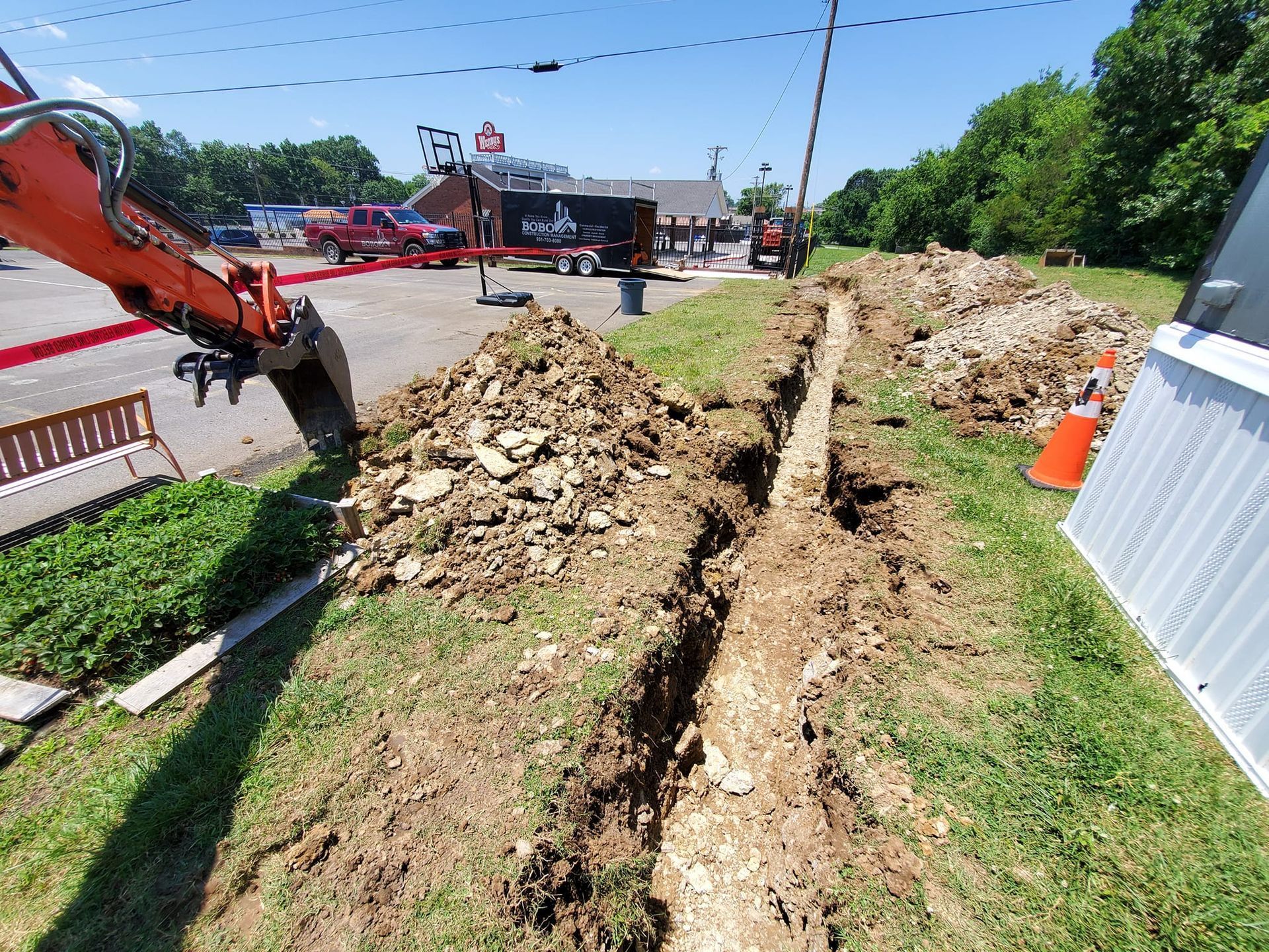 An orange excavator digs a trench along a grassy roadside near a building and parked vehicles under a sunny blue sky.