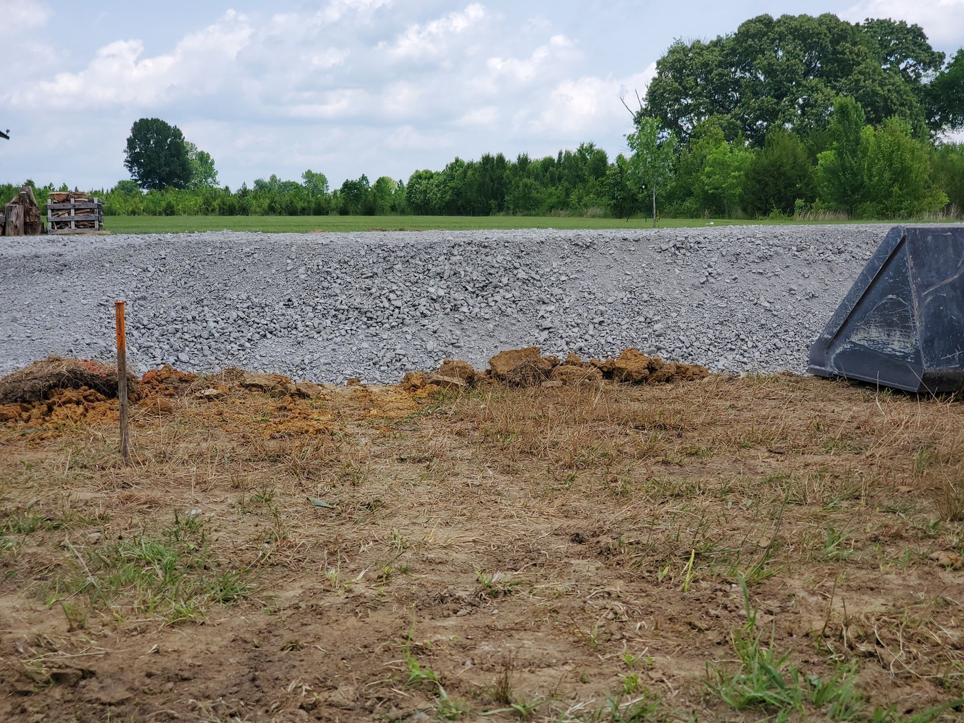 A pile of gray crushed stone sits on a dirt lot, with a tractor bucket visible on the right and green trees in the back.