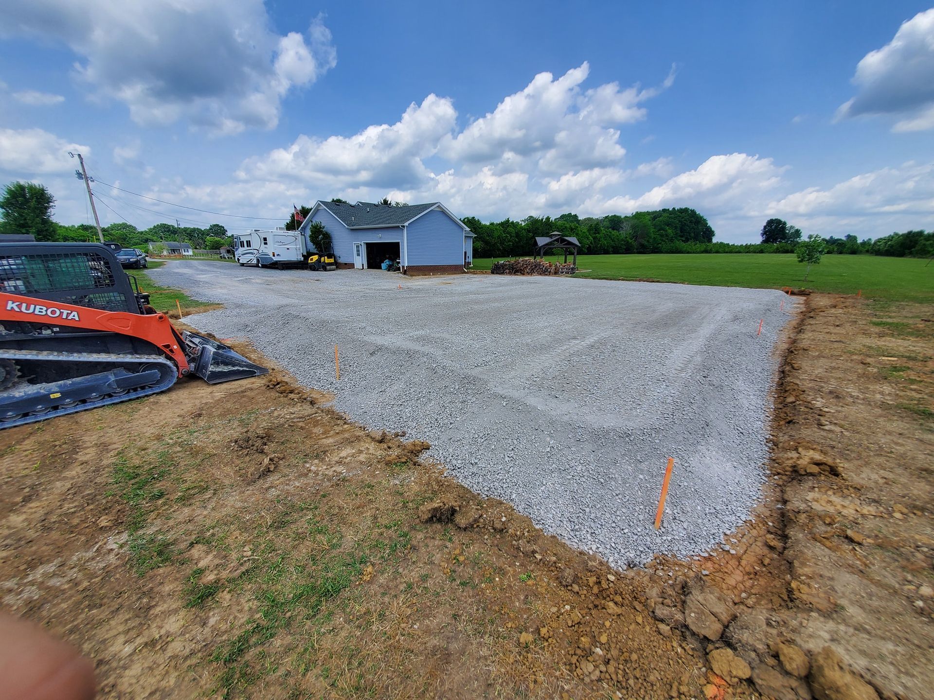 A construction site features a freshly graveled driveway area with a Kubota skid steer parked on the left under a blue sky.
