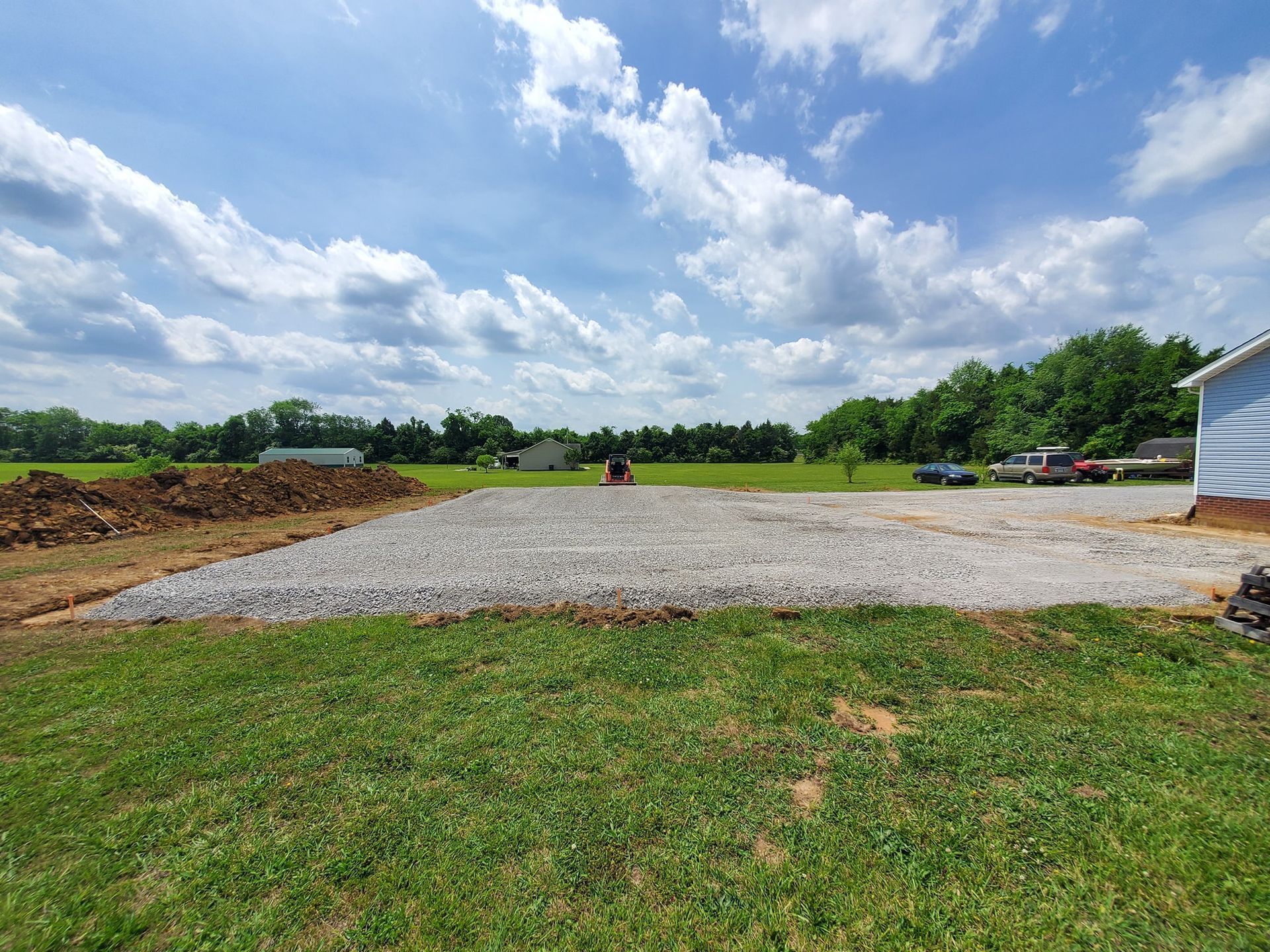 A gravel pad sits in a grassy yard under a blue sky with scattered clouds, next to a building edge and a mound of dirt.