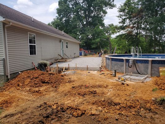 A construction site showing a new concrete patio poured beside a house with a grey vinyl-sided wall and an outdoor pool.