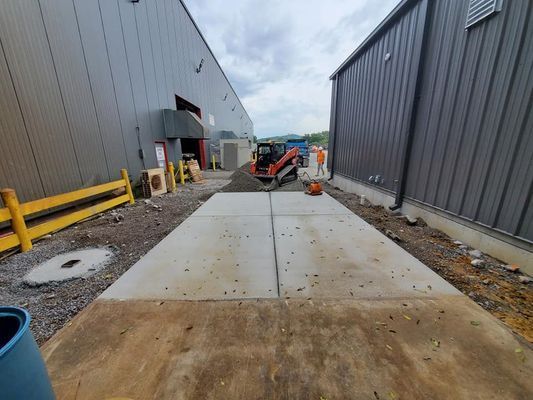 A construction site between two industrial buildings with a newly poured concrete path and a skid-steer loader parked.