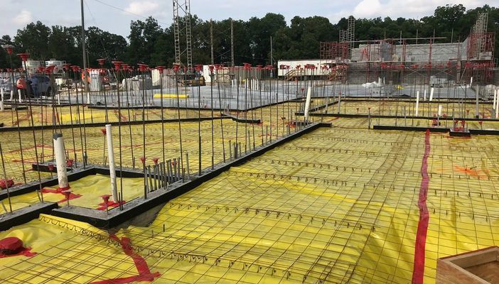 Construction site showing a concrete foundation prepared with yellow vapor barrier, wire mesh, and metal rebar columns.