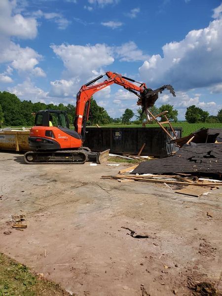 An orange excavator loading demolition debris into a large black dumpster in a grassy, outdoor setting.