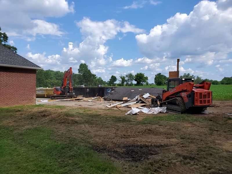 Construction site with an orange excavator and a skid steer loader near a brick building and piles of demolition debris.