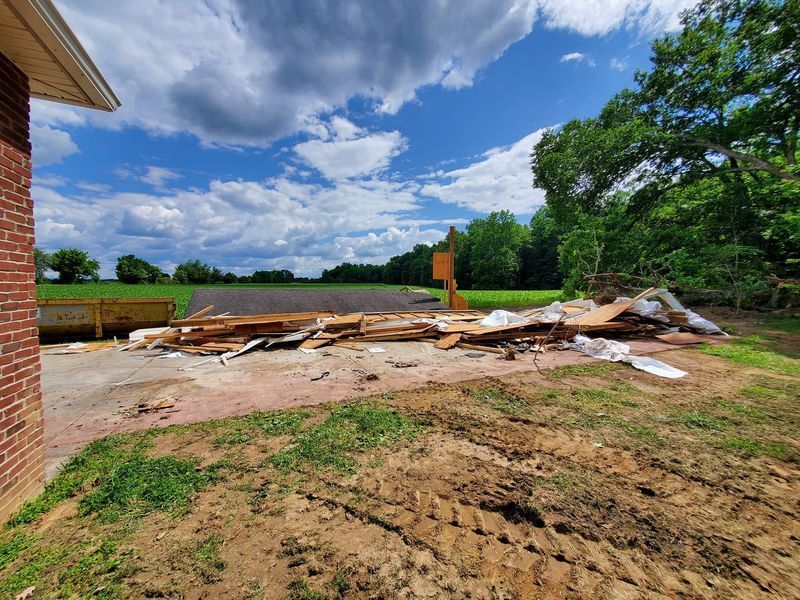 A pile of construction debris and wood planks sits on a concrete slab in a grassy yard under a cloudy blue sky.