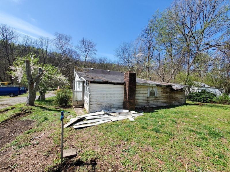 A small, dilapidated white house with a brick chimney stands on a grassy lot under a clear blue sky.