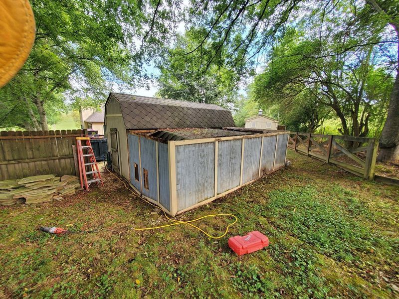 A shed undergoing exterior repairs, showing exposed wooden framing and partial siding in a grassy backyard.