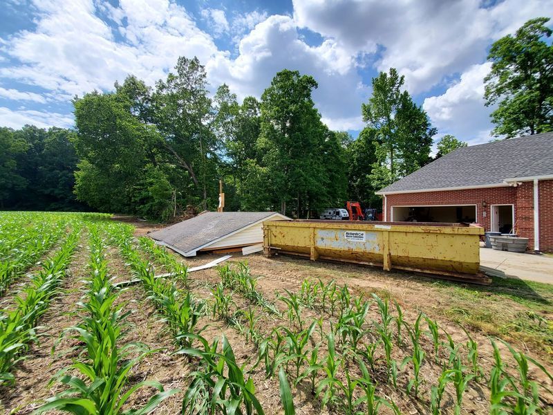 A yellow dumpster sits on a dirt path next to a cornfield and a brick building, with a storm shelter in the background.