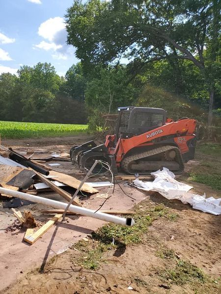An orange Kubota track loader sits in a dirt clearing next to a pile of construction debris and a field.